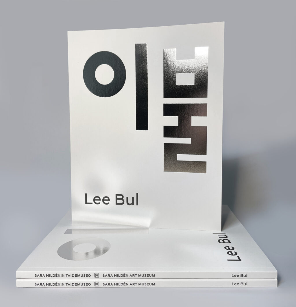 Valkoisia kirjoja pinossa harmaata taustaa vasten. Kirjan kannessa on isoin koreankielisin aakkosin hopealla tekstillä taiteilijan nimi Lee Bul.